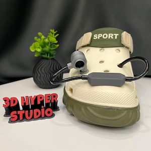 May include: A white and green shoe with the word "SPORT" on the strap. The shoe has a black cord attached to a grey rectangular object. The shoe is sitting on a white surface.