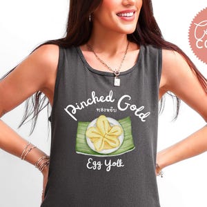 Könnte beinhalten: Dunkelgraues Tanktop mit dem weißen Schriftzug "Pinched Gold Egg Yolk". Die Grafik zeigt ein gelbes Dessert auf einem weißen Teller auf einem grünen Blatt. Das Tanktop gehört zur Comfort Colors Kollektion.