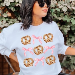Op de afbeelding: Witte sweatshirt met een herhalend patroon van aquarel pretzels en roze strikken. De pretzels zijn goudbruin en de strikken zijn zachtroze. De sweatshirt is een casual, comfortabele stijl.