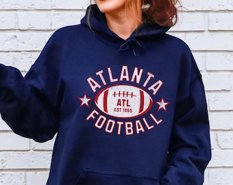 Puede incluir: Sudadera con capucha azul marino con el texto "ATLANTA FOOTBALL" en blanco y rojo, con un gráfico de fútbol americano y el texto "EST 1965". La sudadera tiene un bolsillo delantero y una capucha con cordón.