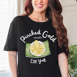 Könnte beinhalten: Schwarzes T-Shirt mit einer Grafik eines gelben Eigelb-Desserts auf einem grünen Blatt, mit den Worten "Pinched Gold" und "Egg Yolk" in weißer Schrift. Das T-Shirt hat einen lässigen Stil.