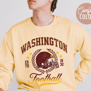 May include: A light yellow sweatshirt with the word "WASHINGTON" in maroon lettering above a maroon football helmet graphic. The word "Football" is written in a script font below the helmet. The sweatshirt has the words "Comfort COLOR" and "Butter" printed on it.