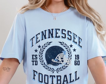 May include: A light blue t-shirt with the words "TENNESSEE FOOTBALL" in a distressed, collegiate-style design. The design includes a football helmet, stars, and a laurel wreath. The shirt is a baby blue color.