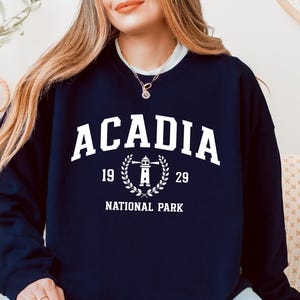 Può includere: Felpa blu navy con la scritta "ACADIA" in lettere bianche. Sotto, una grafica di un faro all'interno di una corona d'alloro, con i numeri "19" e "29" su entrambi i lati. Le parole "NATIONAL PARK" sono stampate sotto.