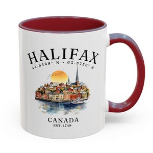 Könnte beinhalten: Ein weißer Keramikbecher mit dunkelrotem Innenraum und Henkel. Die Vorderseite zeigt eine Aquarellillustration der Skyline von Halifax, den Text HALIFAX, die Koordinaten 44.6488 N 63.5752 W, CANADA und EST. 1749 in schwarzer Schrift.