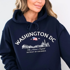 May include: Navy blue hooded sweatshirt featuring the text "WASHINGTON DC" in white, along with a small American flag and a graphic of Washington D.C. landmarks. The text "The Nation's Capital DISTRICT OF COLUMBIA" is also included.