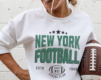 May include: White sweatshirt with the text "NEW YORK FOOTBALL" in green, stars, a football graphic, and "ESTD 1960". A brown football with white laces is held in the image.
