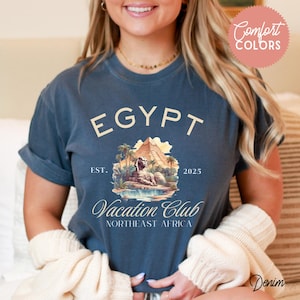 May include: A blue t-shirt with a vintage-style graphic design featuring a sphinx, pyramids, and palm trees. The text on the shirt reads "EGYPT Vacation Club Northeast Africa EST. 2025".