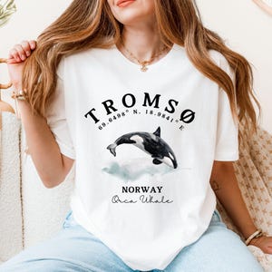 May include: White t-shirt featuring a black graphic of an orca whale leaping from the water. The graphic also includes the text "TROMSO 69.6498° N. 18.9841° E NORWAY Orca Whale".
