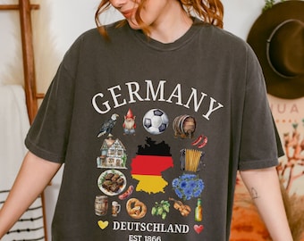 May include: A dark gray t-shirt with a white graphic design featuring the word "GERMANY" in a circle around various images of German culture, including a soccer ball, a pretzel, a beer stein, and a gnome. The text "DEUTSCHLAND EST 1866" is printed below the graphic.