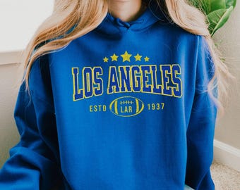 May include: A royal blue hoodie with a yellow and white graphic print that says "Los Angeles" with a football and the text "Esto 1937".