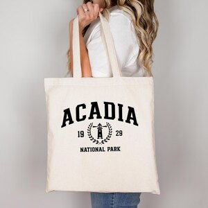 Può includere: Borsa tote color crema con testo nero "ACADIA 1929 NATIONAL PARK". Il design include un faro all'interno di una corona d'alloro. La borsa ha due manici ed è tenuta da una persona.