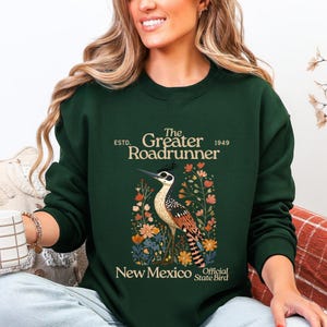 May include: Dark green sweatshirt with a detailed illustration of a roadrunner bird and floral elements. Text includes "The Greater Roadrunner" and "New Mexico Official State Bird". A casual, comfortable style of sweatshirt.