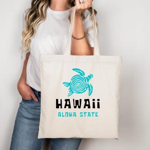 Puede incluir: Un bolso de tela color crema con un gráfico de una tortuga marina turquesa con un patrón de espiral en el caparazón. Debajo de la tortuga, el texto dice HAWAII en letras negras negritas, seguido de las palabras ALOHA STATE en letras mayúsculas turquesas.