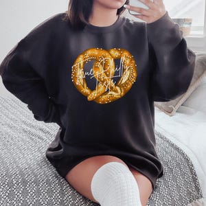 Op de afbeelding: Donkergrijze sweatshirt met een grote pretzel-afbeelding en de tekst "True Philly Taste" in witte letters. De pretzel is goudbruin met witte zoutkorrels. De sweatshirt heeft een casual stijl.