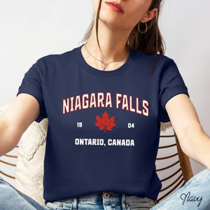 May include: A navy blue t-shirt with white text that reads "NIAGARA FALLS 19 04 ONTARIO, CANADA". A red maple leaf is centred between the years.