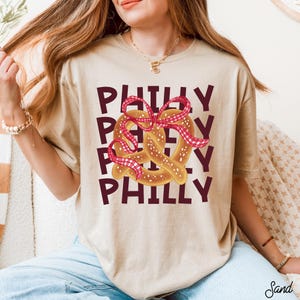 Op de afbeelding: Zandkleurig T-shirt met een pretzel-afbeelding versierd met een rood-wit geruit lint. Het woord "PHILLY" is in een gestapeld ontwerp in donkerrood gedrukt. Een casual kledingstuk voor dagelijks gebruik.