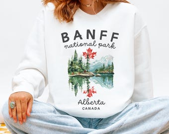 May include: White sweatshirt featuring a scenic illustration of Banff National Park. The design includes a lake, mountains, trees, and a red maple leaf. The text "BANFF national park" and "Alberta CANADA" are printed above and below the illustration.