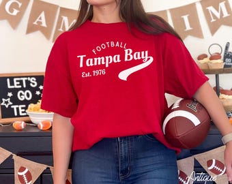 Puede incluir: Una camiseta roja con el texto "FOOTBALL Tampa Bay Est. 1976" en blanco. La camiseta se combina con unos vaqueros azules. En la imagen se sostiene un balón de fútbol. Las palabras "Antique Cherry Red" están escritas en la parte inferior de la imagen.