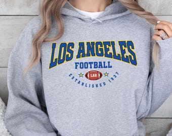 May include: A light grey hoodie featuring the text "LOS ANGELES FOOTBALL" in blue and yellow arched lettering. Below is a football graphic with "LAR" inside. The text "ESTABLISHED 1937" is arched below the football graphic.