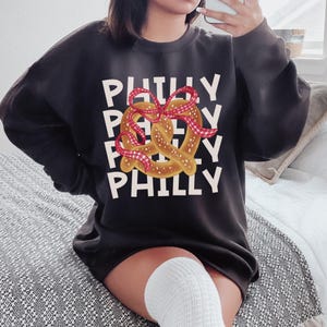 Op de afbeelding: Donkergrijze sweatshirt met het woord "PHILLY" in witte blokletters. Een pretzel met een rood-wit geruit lint staat in het midden. De sweatshirt heeft een casual stijl.