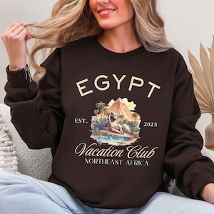 May include: A brown sweatshirt with a graphic print of a sphinx in front of a pyramid. The graphic also includes palm trees, a body of water, and the text "EGYPT EST. 2025 Vacation Club NORTHEAST AFRICA".