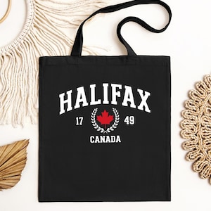 Può includere: Una borsa tote nera con la scritta bianca HALIFAX in carattere arcuato, i numeri 17 e 49 ai lati di una foglia d'acero rossa racchiusa in una corona d'alloro bianca, e la parola CANADA stampata in bianco sotto.