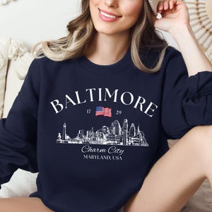 Może przedstawiać: Granatowa bluza z białym nadrukiem przedstawiającym panoramę Baltimore, napisami "Baltimore" i "Charm City, Maryland, USA" oraz małą flagą amerykańską.