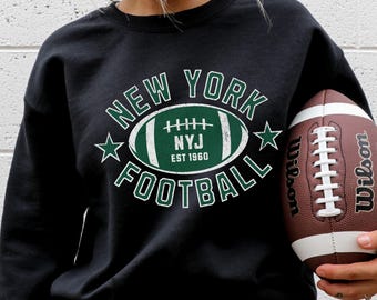 May include: Black sweatshirt with a green and white graphic reading "NEW YORK FOOTBALL" around a football shape with "NYJ EST 1960" inside. A brown and white Wilson football is held.