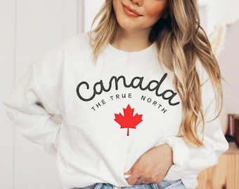 May include: White sweatshirt with the word "Canada" in a script font, the phrase "The True North," and a red maple leaf graphic. The sweatshirt is a long-sleeved crew neck.