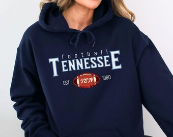 May include: Navy blue hoodie with the word "Tennessee" in white block letters. The word "football" is above the word "Tennessee". A red football graphic with "TNM 1960" is below the word "Tennessee".