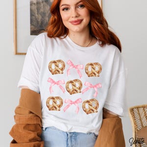 Op de afbeelding: Wit T-shirt met een herhaald patroon van aquarel pretzels en roze strikken. De pretzels zijn goudbruin en de strikken zachtroze. Een casual, alledaagse stijl top.