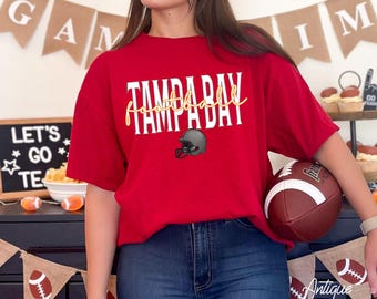 Puede incluir: Una camiseta roja con el texto "TAMPA BAY FOOTBALL DAY" en blanco y dorado, junto con un gráfico de casco de fútbol gris. La persona sostiene un balón de fútbol marrón y blanco. Las palabras "Antique Cherry Red" están escritas en la parte inferior.