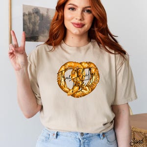 Op de afbeelding: Beige t-shirt met een gedetailleerde afbeelding van een pretzel. De pretzel is goudbruin met zout en de tekst "True Philly Taste" in zwarte letters. Het t-shirt heeft een neutrale kleur.