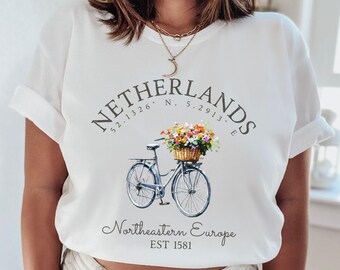May include: White t-shirt featuring a graphic design of a bicycle with flowers in a basket. The text "Netherlands 52.1326. N, 5.2913. E Northeastern Europe EST 1581" is printed on the shirt.
