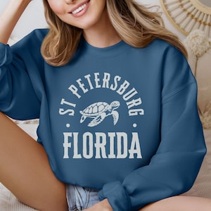 May include: A teal sweatshirt featuring the text "ST. PETERSBURG FLORIDA" in a white, arched font, with a turtle graphic in the center. The sweatshirt is a solid color and has a crew neck.