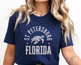 Może przedstawiać: Granatowa koszulka z napisem "ST PETERSBURG FLORIDA" w białym, łukowatym kroju pisma i grafiką żółwia. Koszulka ma okrągły dekolt.