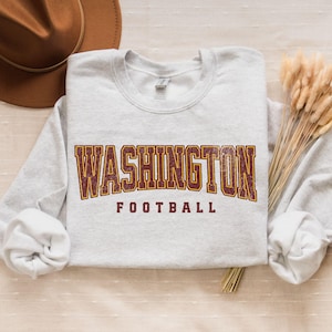 May include: A gray sweatshirt with the text "WASHINGTON FOOTBALL" in maroon and gold letters. The letters have a distressed, vintage look.