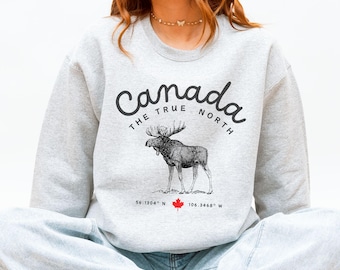 Puede incluir: Sudadera gris claro con la frase "Canada The True North" en fuente cursiva y una ilustración detallada de un alce. Debajo del alce hay coordenadas y una hoja de arce roja.
