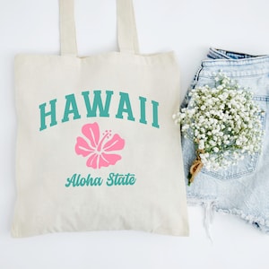 Può includere: Una borsa tote in tela color crema con il testo turchese HAWAII arcuato sopra un grande grafico di un fiore di ibisco rosa, con il testo turchese Aloha State stampato sotto il fiore. La borsa ha due manici e una forma rettangolare.