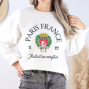 May include: Cream-coloured sweatshirt featuring the text "PARIS FRANCE" in a semi-circular design. The graphic includes a crest with a ship, crown, and laurel wreath. The phrase "Fluctuat nec mergitur" is below the crest.