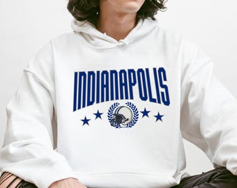 May include: White hooded sweatshirt featuring the word "INDIANAPOLIS" in navy blue block letters. Below the text is a navy blue football helmet graphic with a laurel wreath and four navy blue stars. A casual style sweatshirt.