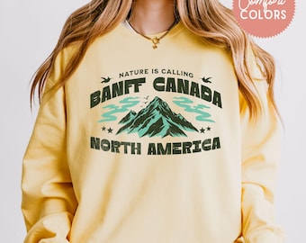 May include: Yellow sweatshirt with a graphic design featuring a mountain range and the text "Nature is calling Banff Canada North America".
