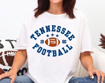 May include: White t-shirt with a blue and orange Tennessee Football graphic. The design includes the word "Tennessee" arched above a football graphic, with the years "19" and "60" on either side. The word "Football" is arched below.