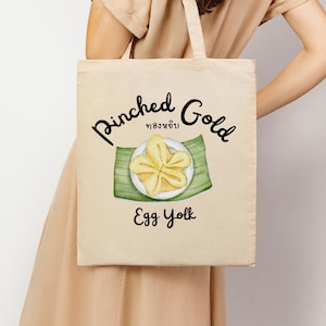 Könnte beinhalten: Beige Tragetasche mit den Worten "Pinched Gold" und "Egg Yolk" in schwarzer Schrift. Die Tasche zeigt eine Aquarellillustration eines gelben, blütenförmigen Desserts auf einem grünen Blatt. Die Tasche hat ein schlichtes Design mit einem Griff.