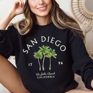 May include: Black sweatshirt with the words "SAN DIEGO" in white arched text above a watercolor illustration of three palm trees. Below the trees are the numbers "17" and "96", and the text "La Jolla Beach CALIFORNIA".