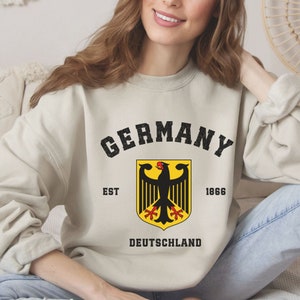 May include: A beige sweatshirt with the word "GERMANY" in black above a yellow and black emblem of a German eagle. The text "EST 1866" and "DEUTSCHLAND" are also printed on the sweatshirt.