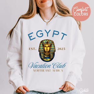 May include: White sweatshirt with the word "EGYPT" in blue block letters above a graphic of a pharaoh's mask. The text "Vacation Club" is below the graphic, with "NORTHEAST AFRICA" underneath. The sweatshirt also has the text "EST. 2025".