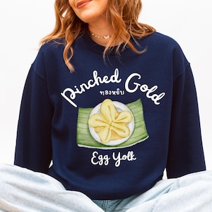 Könnte beinhalten: Marineblaues Sweatshirt mit den Wörtern "Pinched Gold" und "Egg Yolk" in weißer Schrift. Das Design zeigt eine Aquarellillustration eines gelben Eigelbgerichts auf einem grünen Blatt.