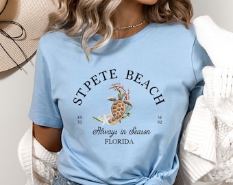 Może przedstawiać: Jasnoniebieski t-shirt z grafiką przedstawiającą żółwia morskiego, koralowiec i rozgwiazdę. Napis głosi ST. PETE BEACH, ESTD 1892, Always in Season oraz FLORIDA czarnymi literami. Koszulka ma krótkie rękawy i okrągły dekolt.
